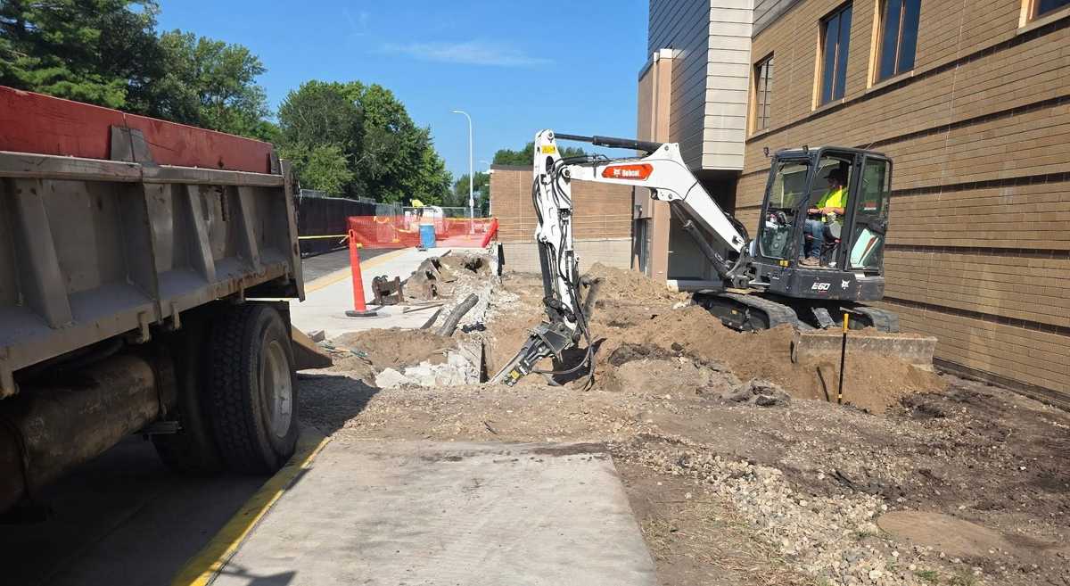 Compact excavator with a hydraulic hammer removing concrete beside a brick building, with a dump truck in the foreground