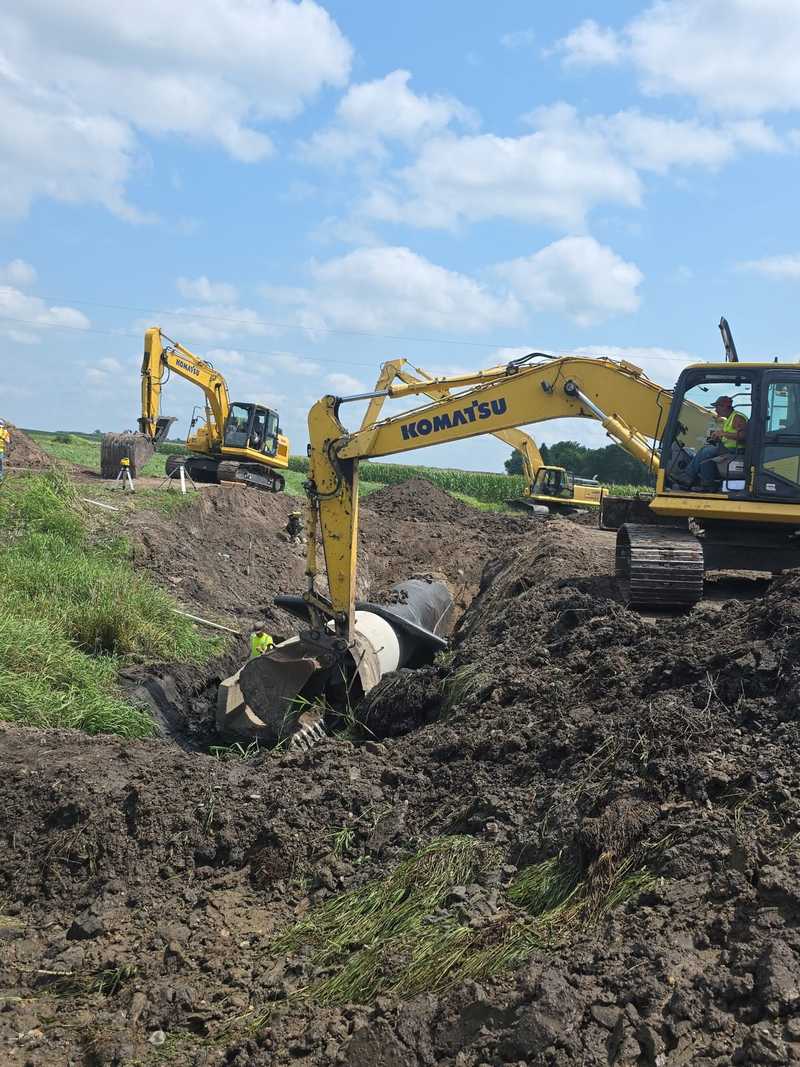 Two excavators working in a freshly dug ditch beside a farm field, one with a large pipe at its bucket