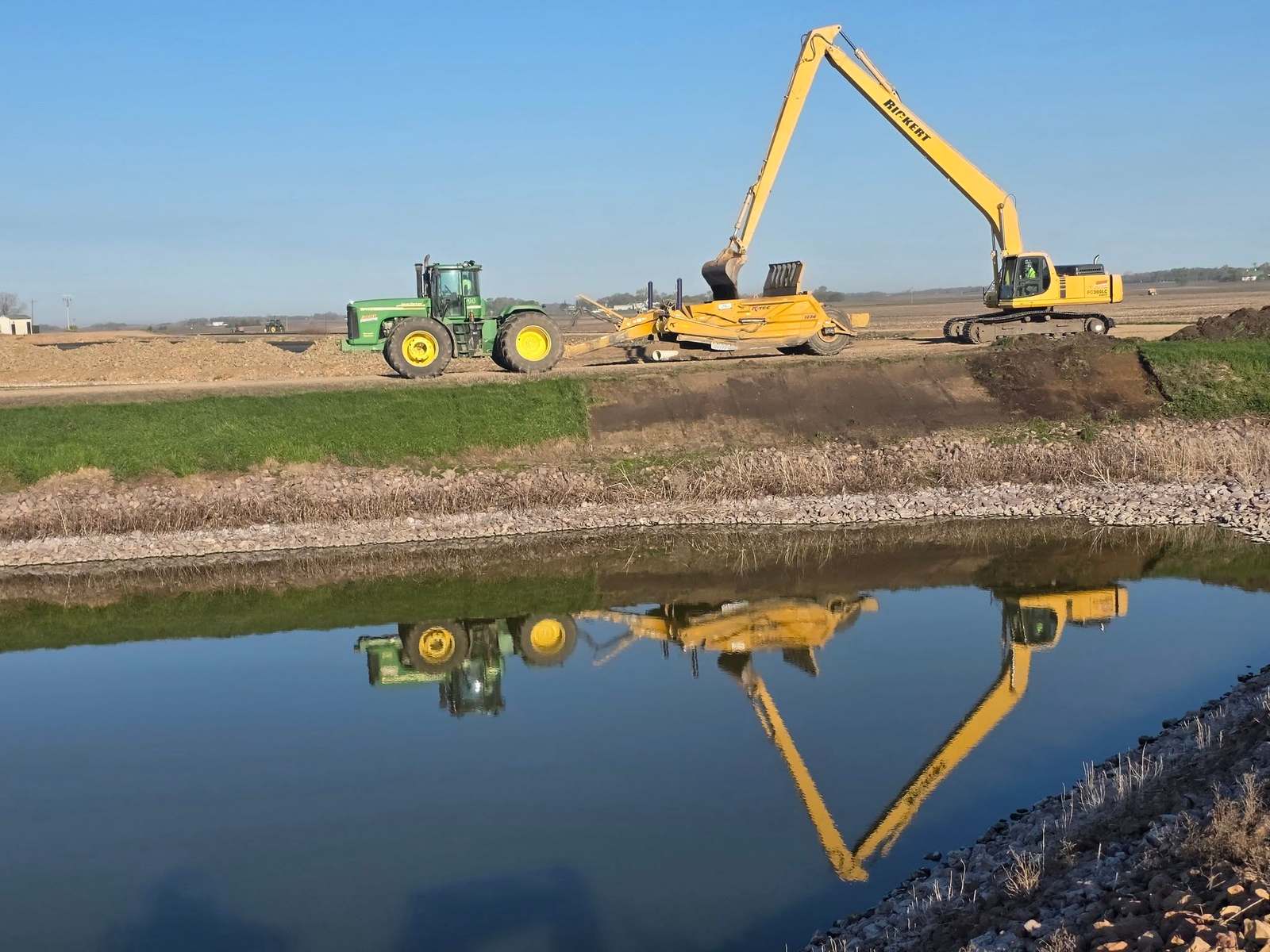 Long-reach excavator and a tractor with a scraper working beside open water under a clear sky