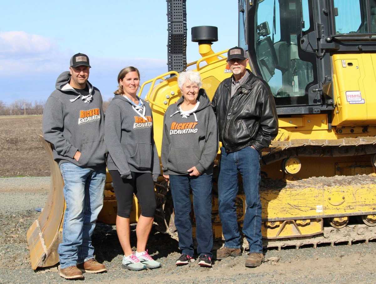 Four people wearing Rickert Excavating sweatshirts standing in front of a yellow bulldozer