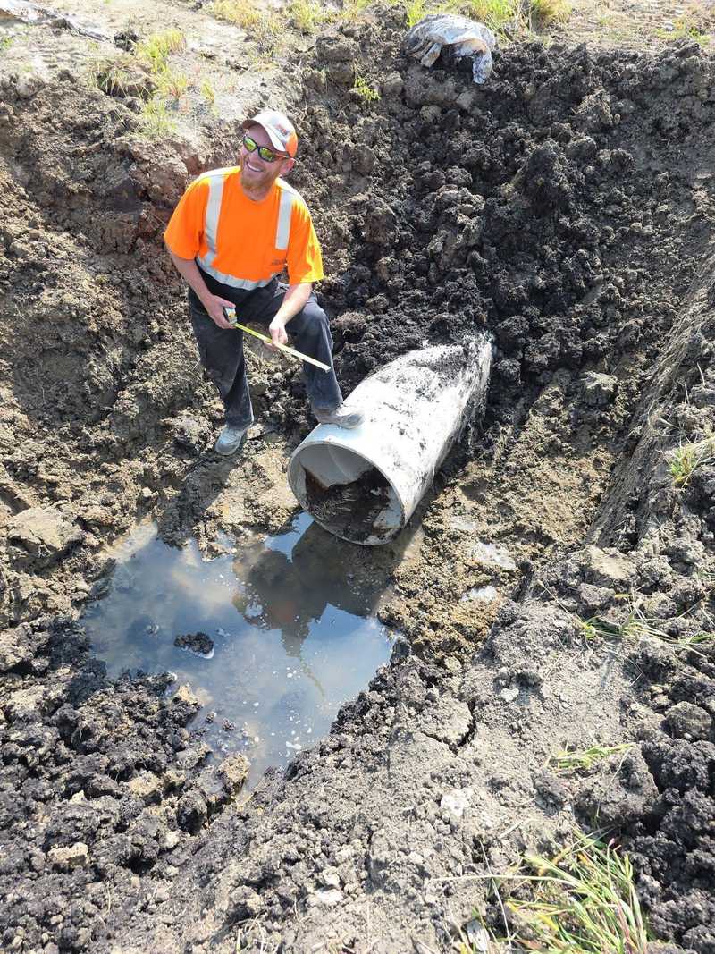 Worker in an orange safety shirt crouching in a deep trench next to a large concrete pipe